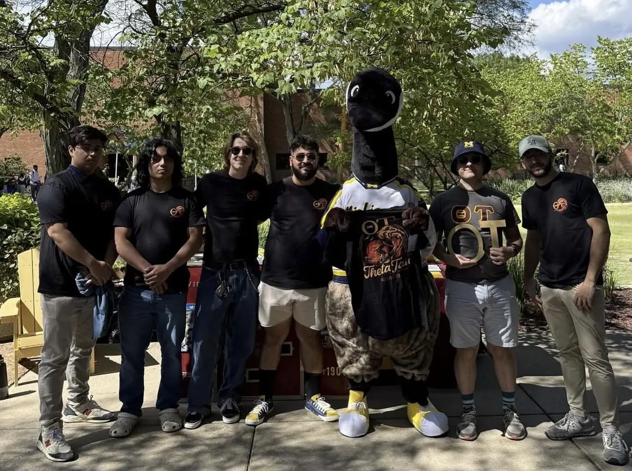 Brothers in chapter shirts with the UM-Dearborn goose mascot at an outdoor campus event.
